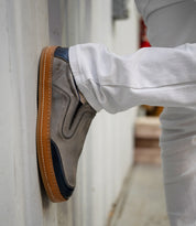 Wearing crisp white pants, the person showcases the timeless silhouette of Bed|Stü’s Lanius slip-on shoes—crafted in gray suede with navy accents and gum soles—as they rest their foot against a wall.