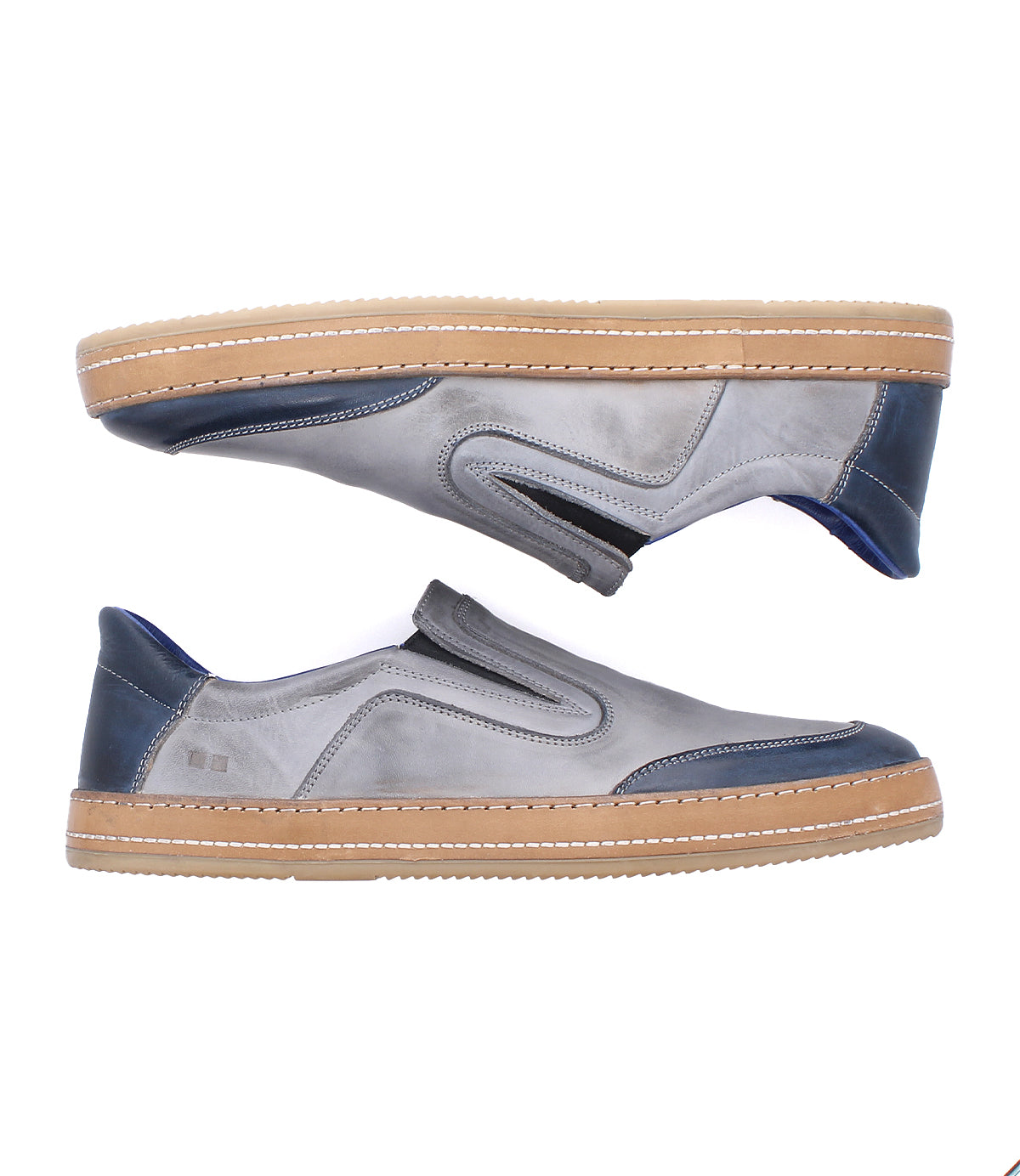 The Lanius by Bed Stu features a timeless silhouette in gray with blue accents and tan rubber soles. Shown from the side, one shoe faces up while the other faces down, highlighting modern color blocking and slip-on design.