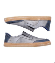 The Lanius by Bed Stu features a timeless silhouette in gray with blue accents and tan rubber soles. Shown from the side, one shoe faces up while the other faces down, highlighting modern color blocking and slip-on design.