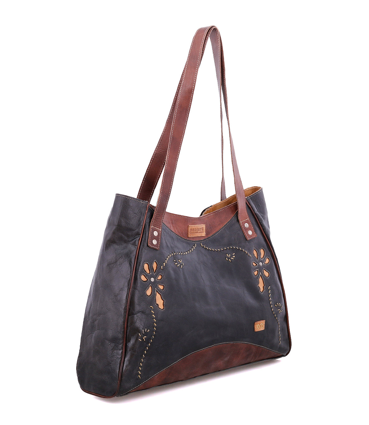The Larue LTC by Bed|Stü is a black and brown leather shoulder bag adorned with floral embroidery and elegant floral motif cutouts. It features two long shoulder straps, a small stitched Bed|Stü label on the front, and refined handstitched details for an artisanal finish, making this versatile tote both stylish and distinctive.