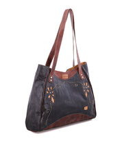 The Larue LTC by Bed|Stü is a black and brown leather shoulder bag adorned with floral embroidery and elegant floral motif cutouts. It features two long shoulder straps, a small stitched Bed|Stü label on the front, and refined handstitched details for an artisanal finish, making this versatile tote both stylish and distinctive.