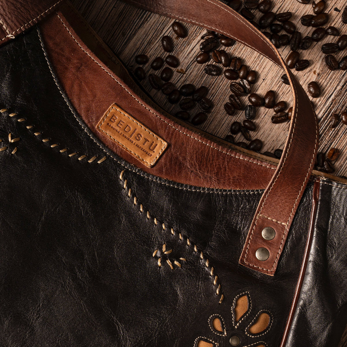 A black and brown Bed|Stü Larue LTC shoulder bag with handstitched details and a floral motif cutout, displayed on a wooden surface with scattered coffee beans.