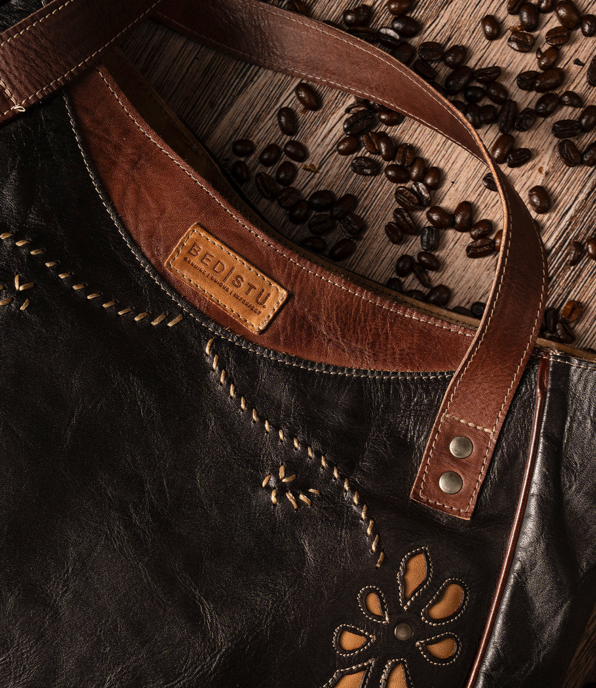 A black and brown Bed|Stü Larue LTC shoulder bag with handstitched details and a floral motif cutout, displayed on a wooden surface with scattered coffee beans.
