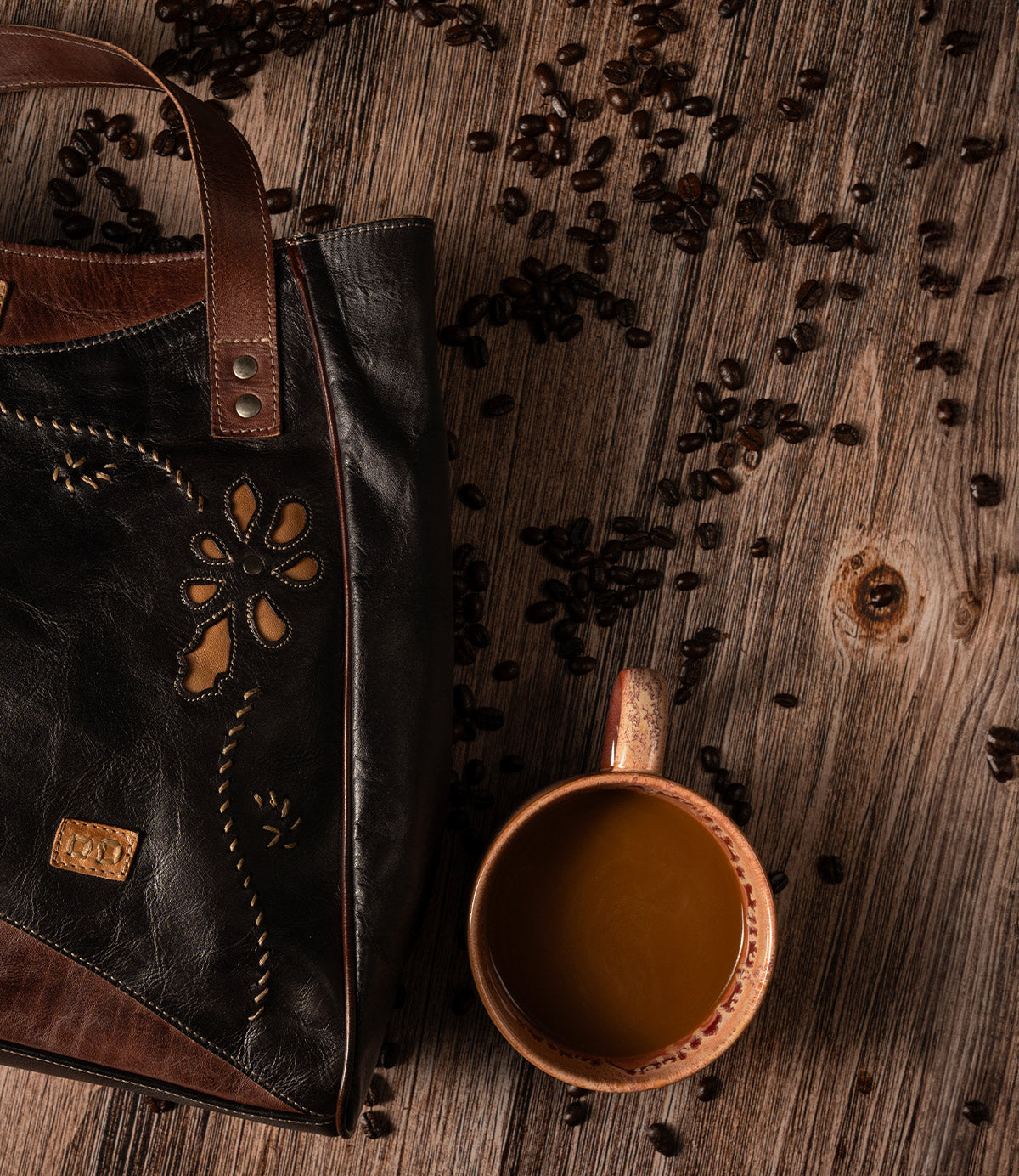 A black and brown leather Larue LTC shoulder bag with floral motif cutouts sits next to a mug of coffee and scattered coffee beans on a wooden surface.