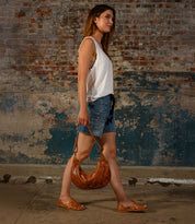 A woman carrying the Bed Stu Lauren II brown handbag walks past a textured brick wall, dressed in a white sleeveless top, denim shorts, and brown huarache sandals.