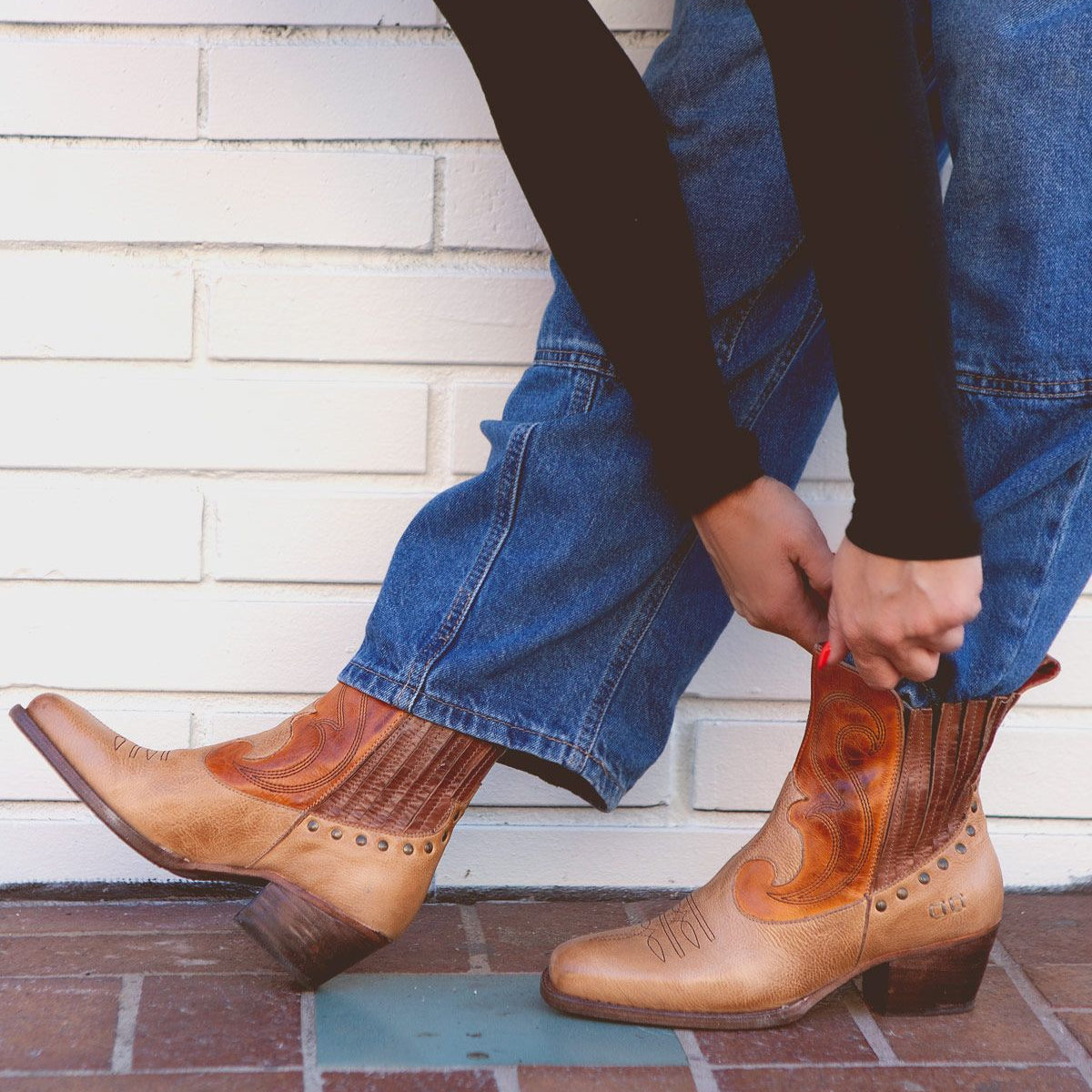 Person wearing blue jeans and a black top, pulling on the Bed Stu League Western short boots with playful stitching details and a low heel.