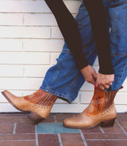 Person wearing blue jeans and a black top, pulling on the Bed Stu League Western short boots with playful stitching details and a low heel.