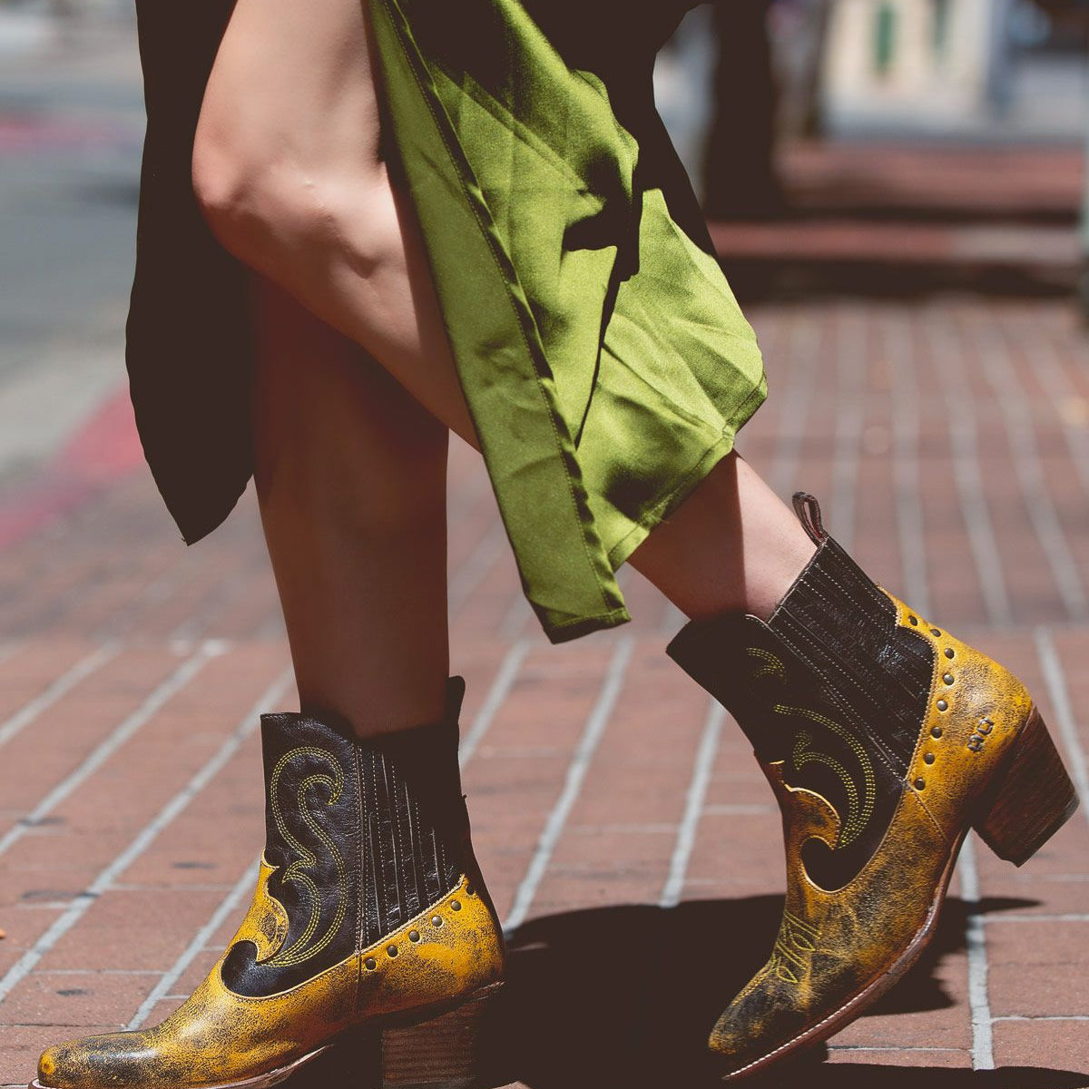 A person wearing yellow and black League boots by Bed Stu walks on a brick sidewalk, with a green skirt billowing around their legs.