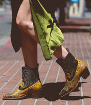 A person wearing yellow and black League boots by Bed Stu walks on a brick sidewalk, with a green skirt billowing around their legs.