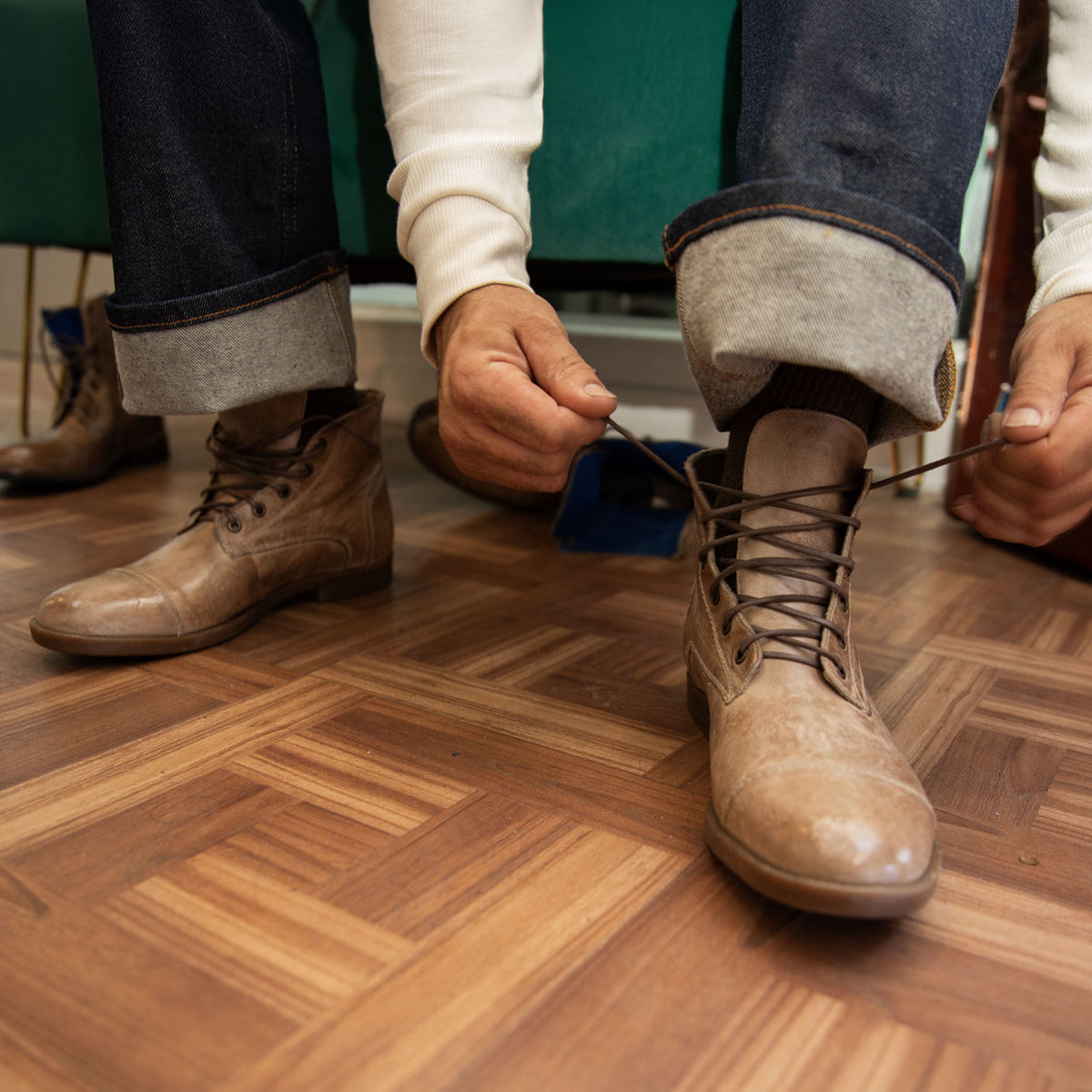 A person wearing jeans and a white sweater ties the laces of Bed Stu's Leonardo brown leather boots, which feature a comfortable insole, on a parquet floor.