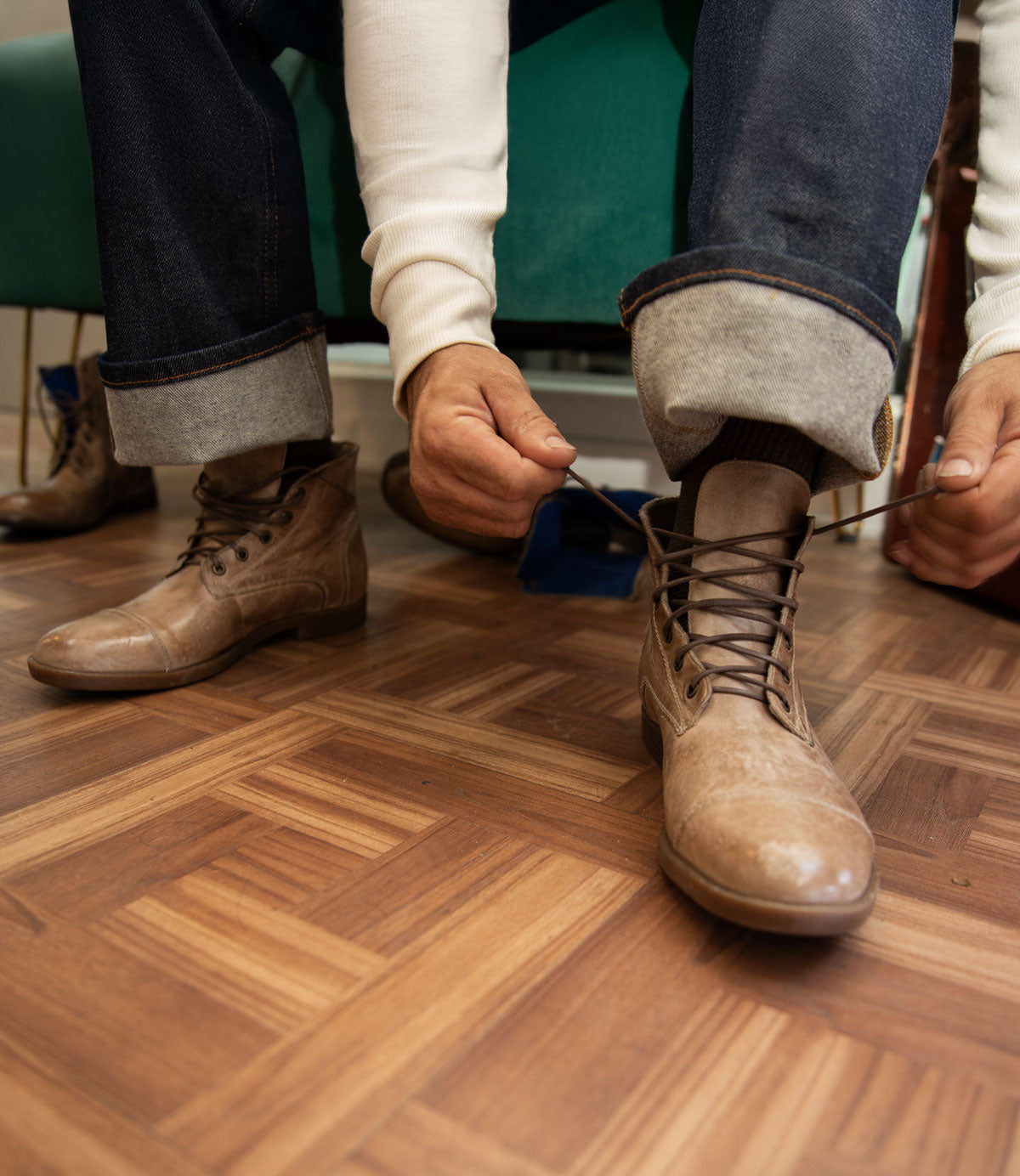 A person wearing jeans and a white sweater ties the laces of Bed Stu's Leonardo brown leather boots, which feature a comfortable insole, on a parquet floor.