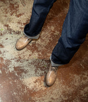 A person wearing the Leonardo shoes by Bed Stu, featuring rolled-up dark jeans and tan dress shoes with a breathable insole, standing on a worn, textured floor.