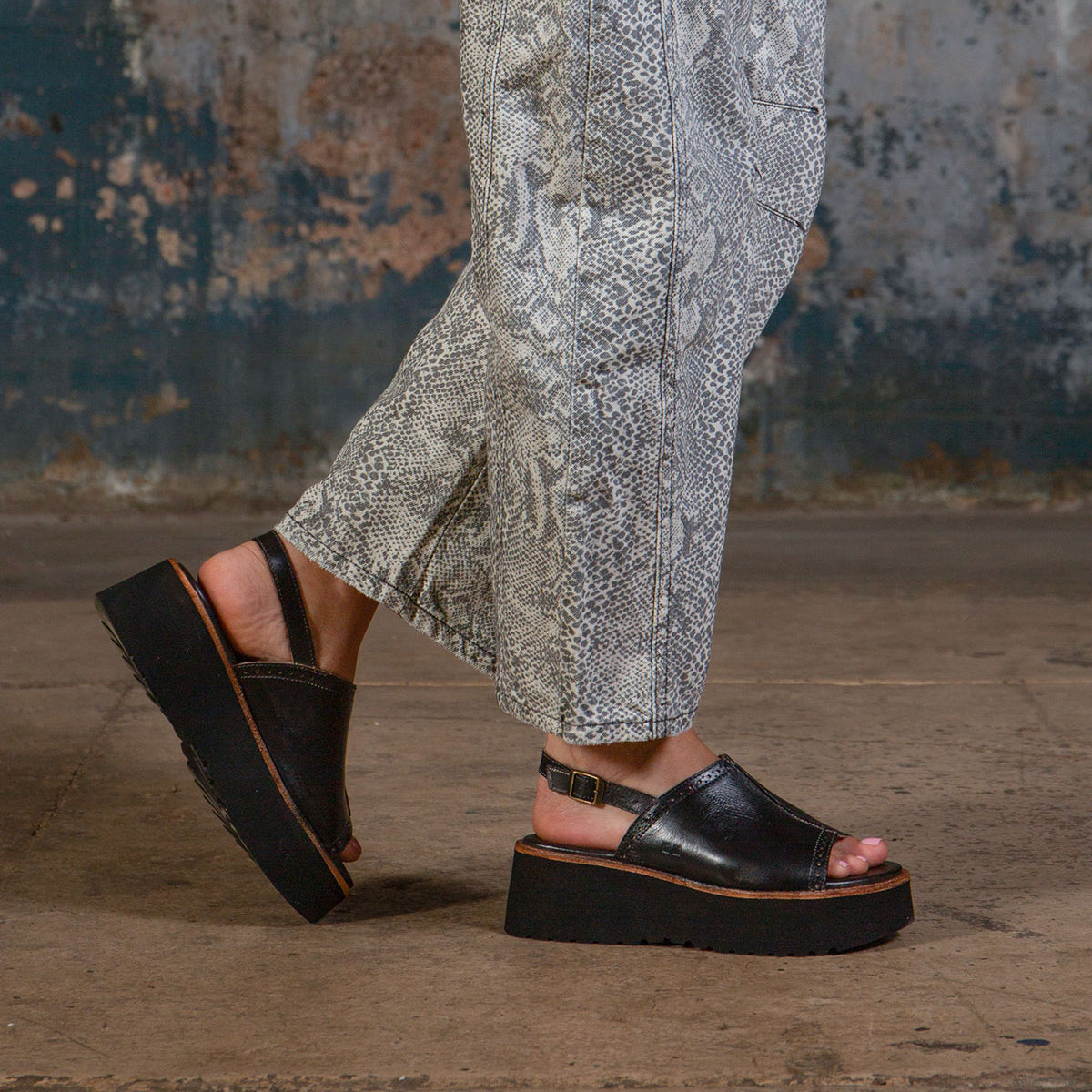 A person in Bed Stu’s Library snakeskin-patterned pants and black platform sandals stands on a concrete floor with a worn, textured wall behind them, channeling retro style.
