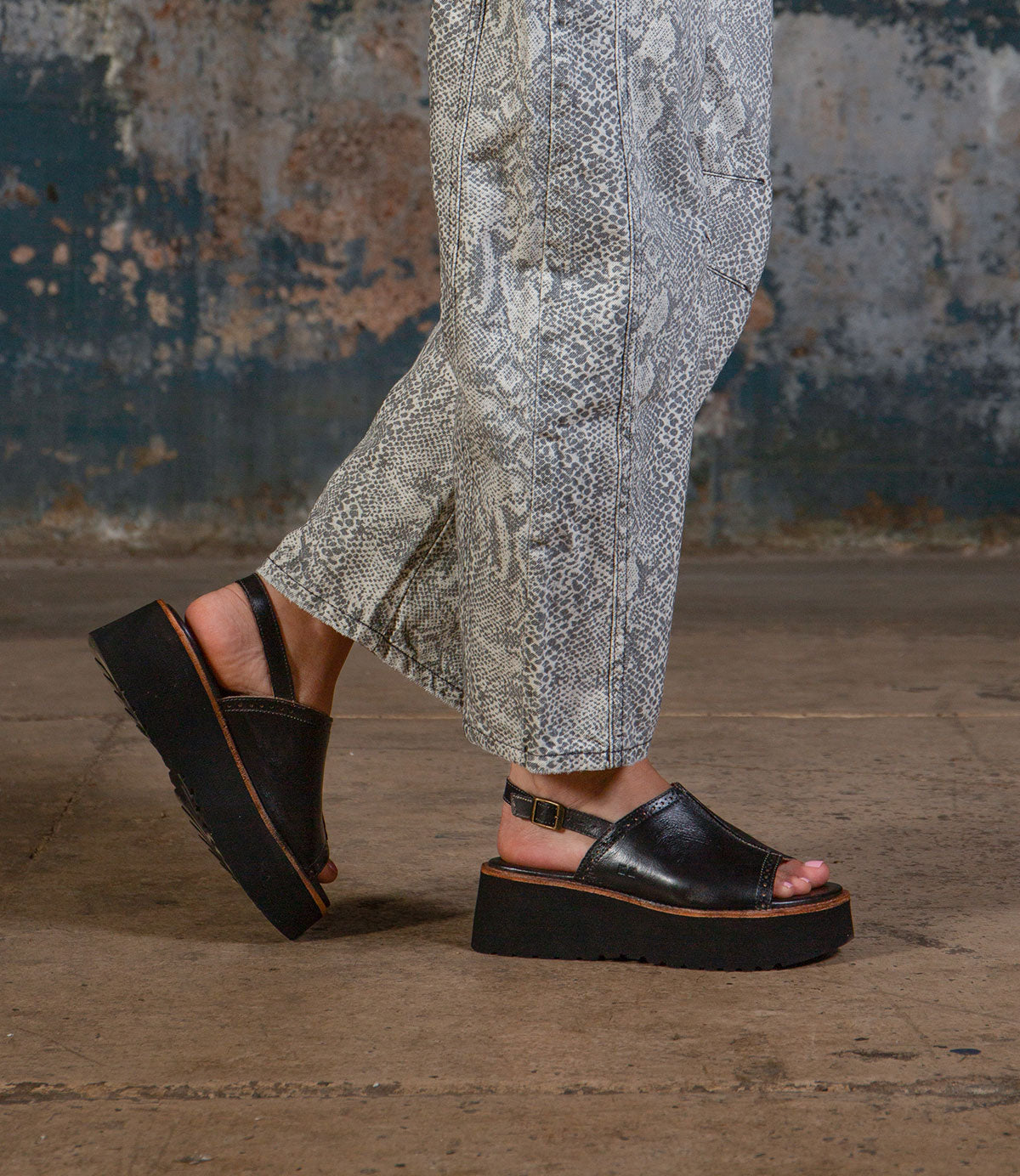 A person in Bed Stu’s Library snakeskin-patterned pants and black platform sandals stands on a concrete floor with a worn, textured wall behind them, channeling retro style.