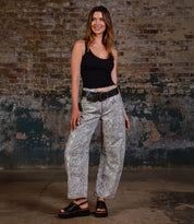 A woman channels a ‘90s vibe indoors against a brick wall, wearing Bed Stu’s Library wide-legged snakeskin print pants, a black tank top, black platform sandals, and a statement black belt with a large buckle.