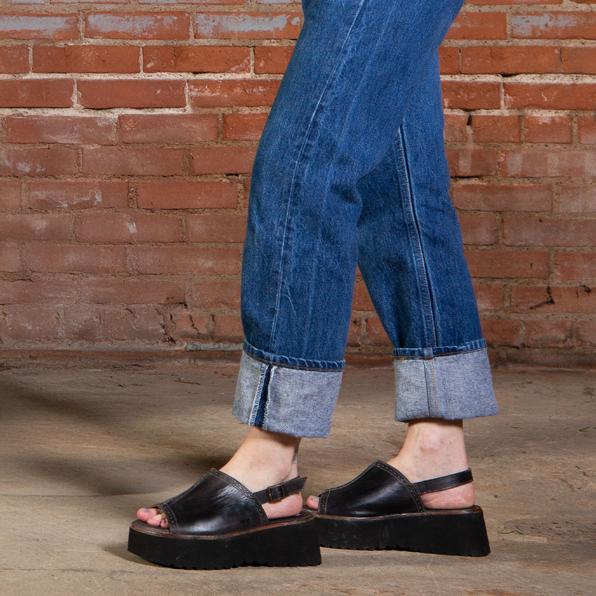 A person in cuffed blue jeans and Bed Stu’s Library black platform sandals stands on a concrete floor with a brick wall behind them, channeling ’90s nostalgia.