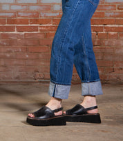 A person in cuffed blue jeans and Bed Stu’s Library black platform sandals stands on a concrete floor with a brick wall behind them, channeling ’90s nostalgia.