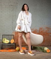 A woman wearing the Bed Stu Library platform sandals smiles indoors beside a modern white chair, with cut melons and metal baskets on the floor, evoking a subtle '90s vibe.