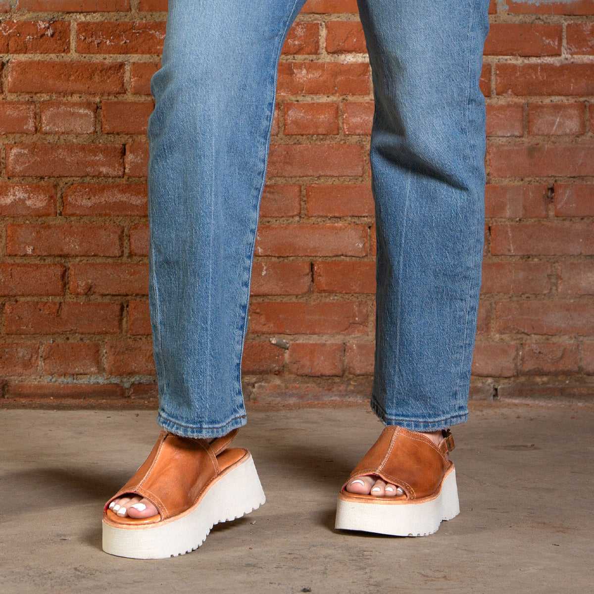 A person wearing blue jeans and Bed Stu's Library retro-style footwear with a chunky platform sole stands on a concrete floor in front of a brick wall.