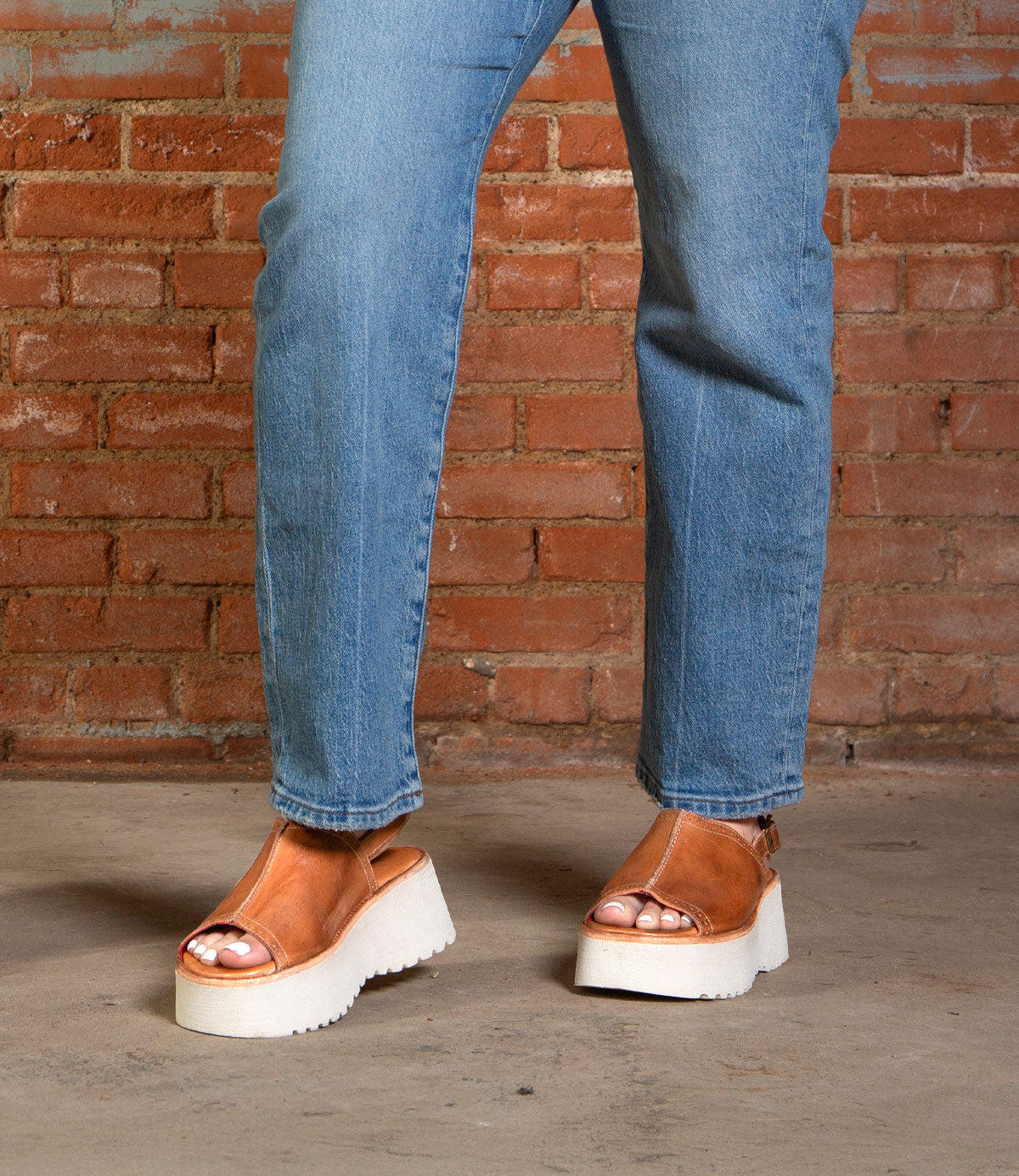 A person wearing blue jeans and Bed Stu's Library retro-style footwear with a chunky platform sole stands on a concrete floor in front of a brick wall.