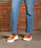 A person wearing blue jeans and Bed Stu's Library retro-style footwear with a chunky platform sole stands on a concrete floor in front of a brick wall.