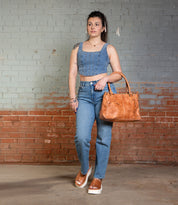 A woman leans against a brick wall in a denim crop top and jeans, channeling '90s vibes with the Bed Stu "Library" large tan handbag and platform sandals.