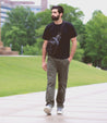 A man with a beard, wearing a black t-shirt, green pants, and vintage Bed Stu Lighthouse low-top sneakers, walks on a paved path in a park on a cloudy day.