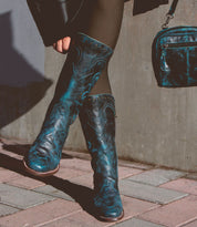 A person wearing Bed Stu's Limit mid-calf Western boots in black and teal patterns with black tights, holding a matching teal purse.