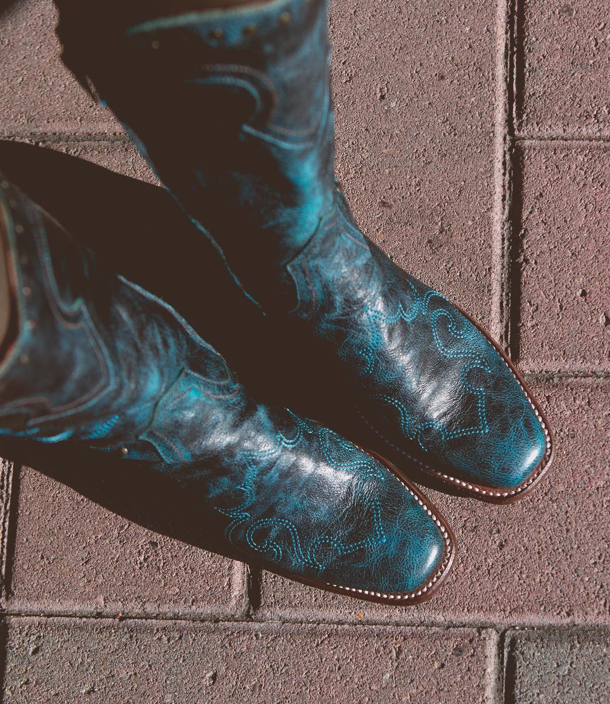 A pair of black Bed Stu Limit Western mid-calf boots with blue floral stitching, seen from above, standing on red brick pavement.