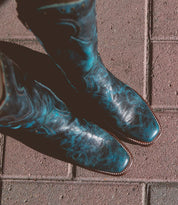 A pair of black Bed Stu Limit Western mid-calf boots with blue floral stitching, seen from above, standing on red brick pavement.