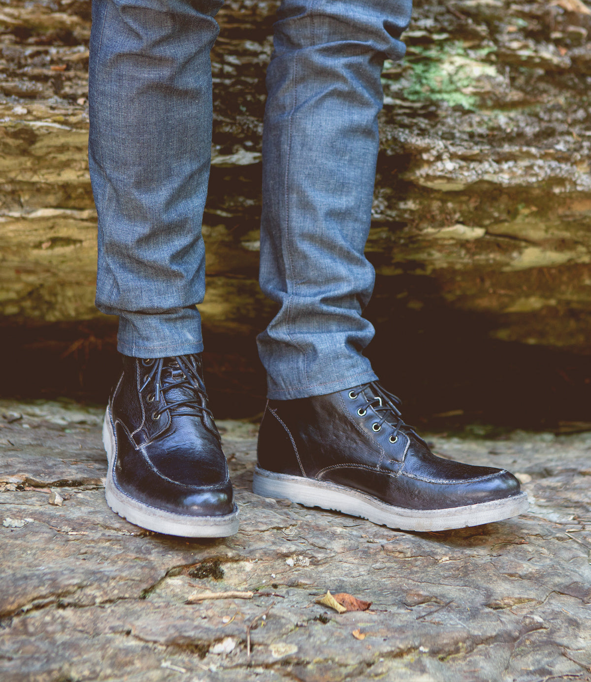 Close-up of a person wearing dark blue Lincoln jeans and Bed Stu black leather work boots standing on a rocky surface.