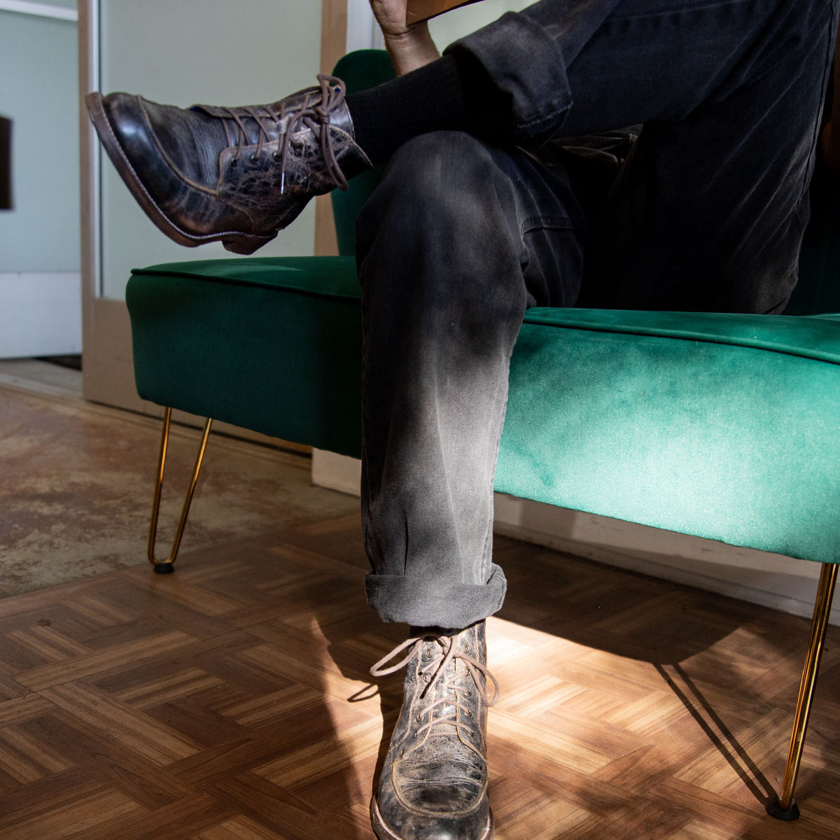 A person sitting on a green sofa with their legs crossed is holding a notebook. They are wearing jeans and the Lincoln II black lace-up boots by Bed Stu. Sunlight shines on the floor, emphasizing the comfort provided by the breathable insoles.