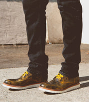 Person wearing black pants and metallic-colored Bed Stu Lincoln boots with yellow laces, standing outdoors on a concrete surface.