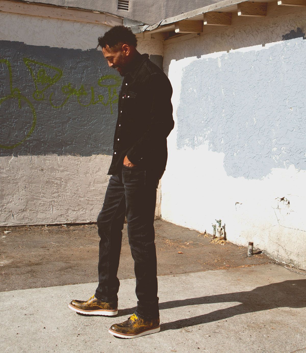 Person in a black shirt and jeans stands on a sidewalk next to a graffitied and worn wall in sunlight, their Bed Stu Lincoln leather boots adding a rugged touch to the scene.