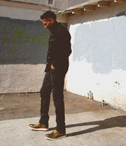 Person in a black shirt and jeans stands on a sidewalk next to a graffitied and worn wall in sunlight, their Bed Stu Lincoln leather boots adding a rugged touch to the scene.