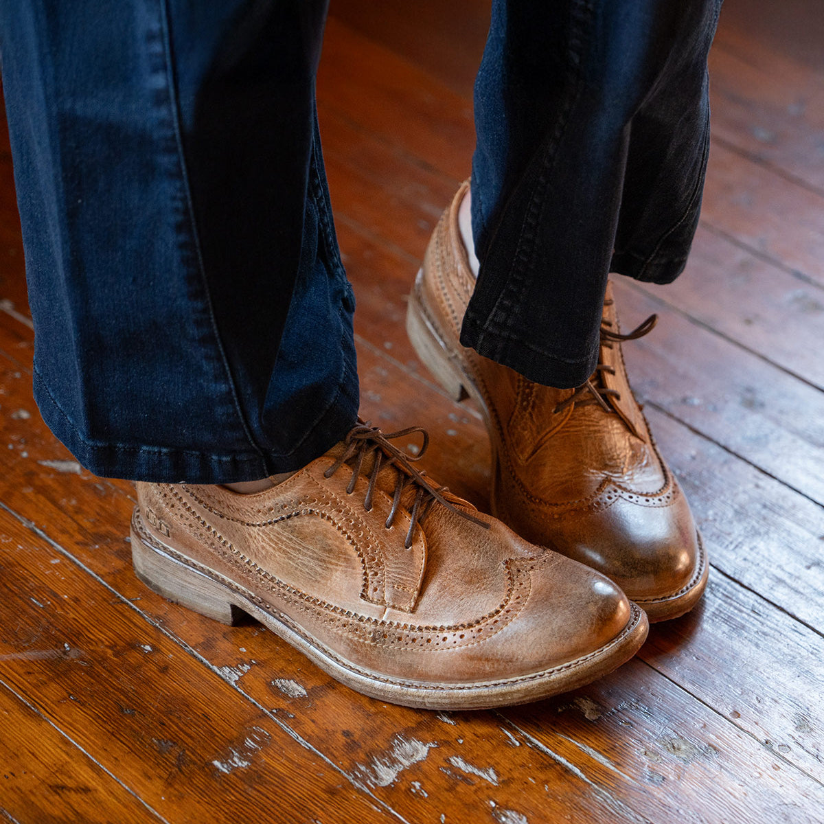Someone stands on a worn wooden floor, wearing dark pants and Bed Stu's Lita K II brown leather wingtip shoes with brogue details.