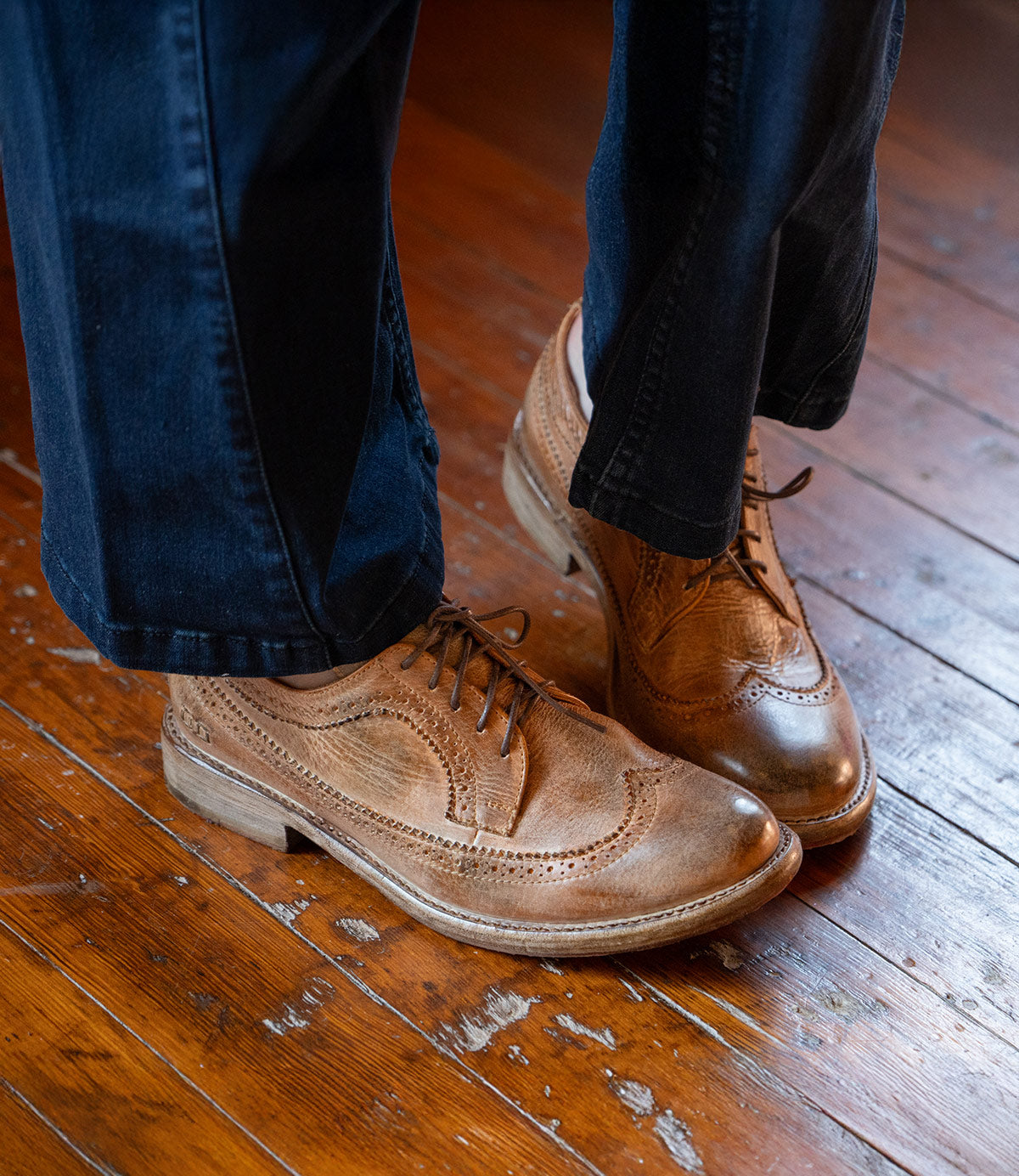 Someone stands on a worn wooden floor, wearing dark pants and Bed Stu's Lita K II brown leather wingtip shoes with brogue details.