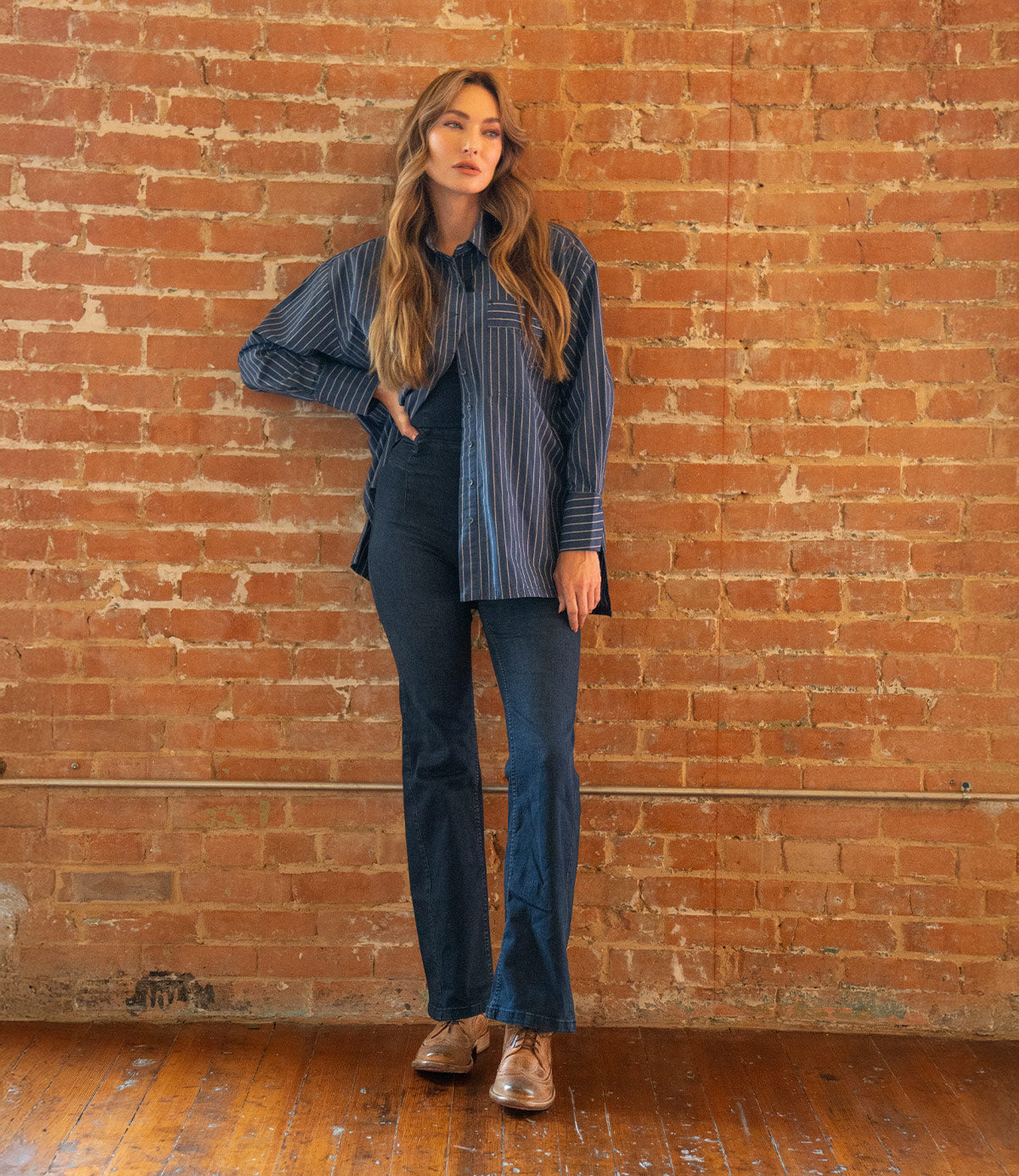 A woman stands indoors against a brick wall, wearing a blue striped oversized shirt, dark blue pants, and Bed Stu's Lita K II brown wingtip shoes with brogue details, confidently resting one hand on her hip.