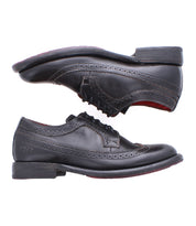 The Bed Stu Lita K II showcases two black leather wingtip shoes with brogue detailing and classic derby laces, displayed horizontally and opposite each other.