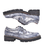 The Lita K III lace-up shoes by Bed Stu, featuring distressed metallic silver leather and chunky black soles that capture the essence of 90’s fashion, are stylishly positioned with one shoe on top of the other.