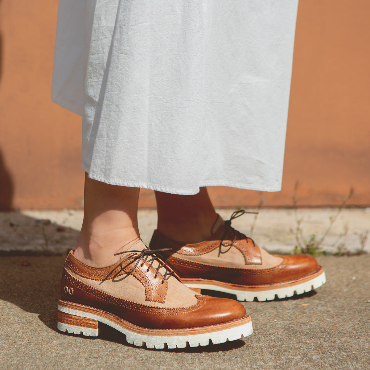 A person standing in Bed Stu Lita K III platform shoes, embodying 90’s fashion, paired with a white skirt against an orange wall.