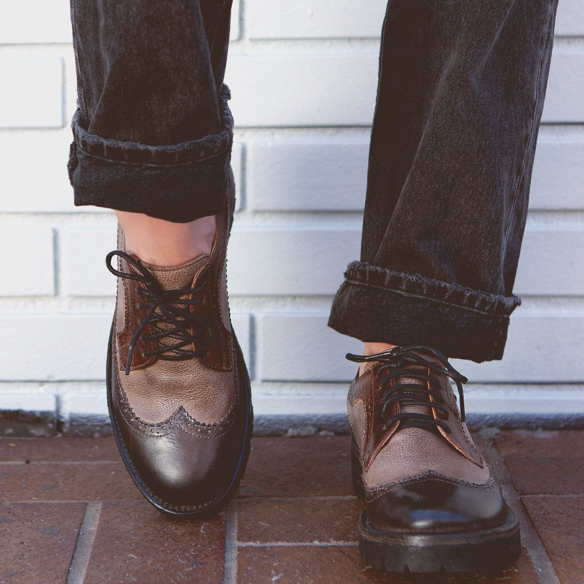 Person wearing black jeans, brown leather wingtip shoes from Bed Stu, standing on a tiled surface in front of a white brick wall.