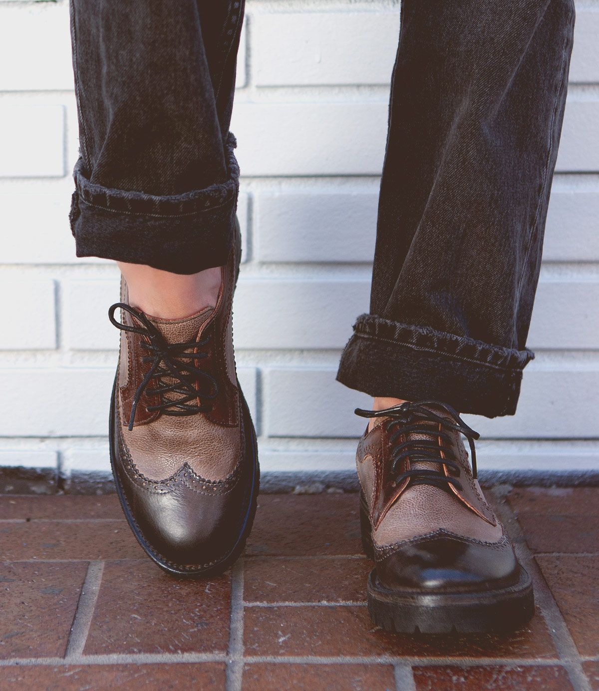 Person wearing black jeans, brown leather wingtip shoes from Bed Stu, standing on a tiled surface in front of a white brick wall.