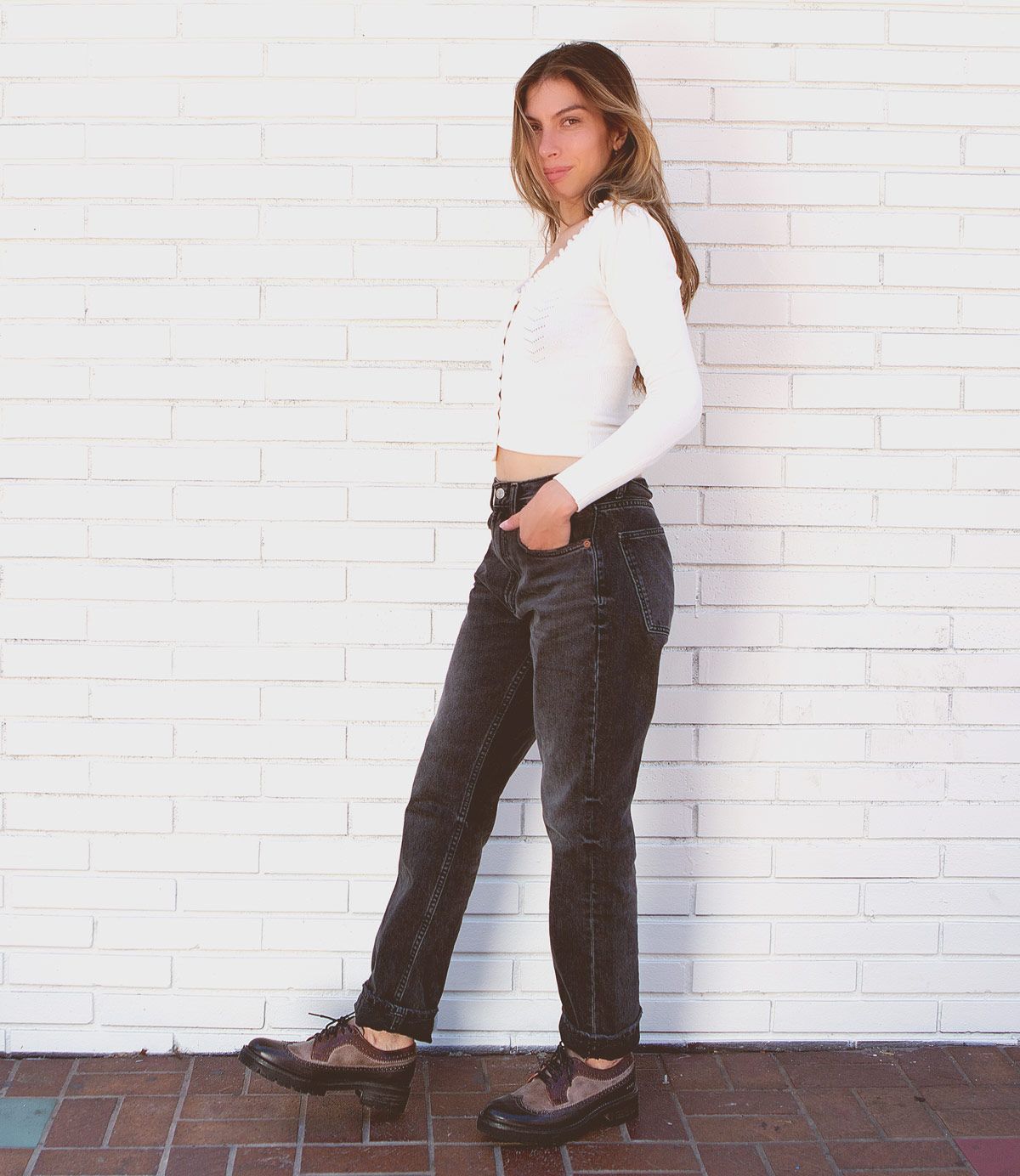 A person stands against a white brick wall, wearing the Proem white long-sleeve top by Bed Stu, dark jeans with a vintage metal belt buckle, and black shoes. Their hands are in their pockets as they look at the camera.