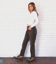 A person stands against a white brick wall, wearing the Proem white long-sleeve top by Bed Stu, dark jeans with a vintage metal belt buckle, and black shoes. Their hands are in their pockets as they look at the camera.