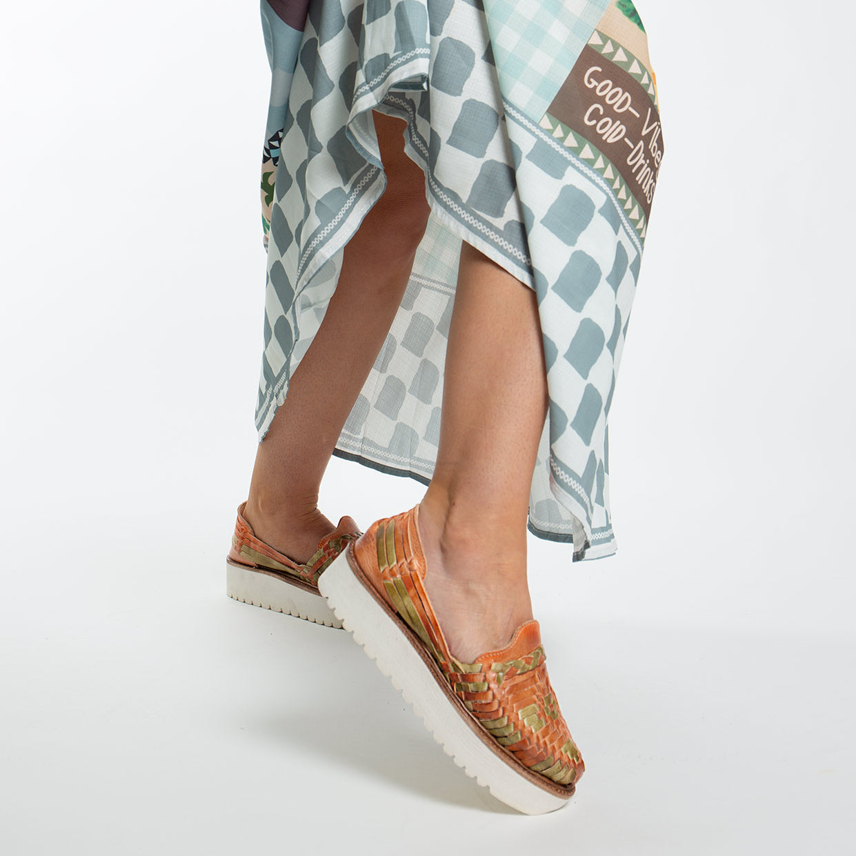 A person strides confidently in Bed Stu's Lizzie orange woven slip-on shoes with cushy XL EXTRALIGHT® outsoles. Paired with a blue and white text-adorned checkered skirt, each step is full of flair.