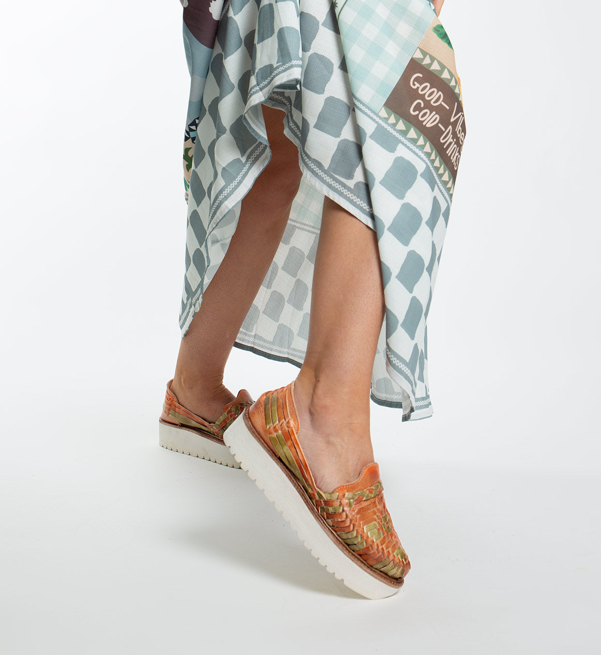 A person strides confidently in Bed Stu's Lizzie orange woven slip-on shoes with cushy XL EXTRALIGHT® outsoles. Paired with a blue and white text-adorned checkered skirt, each step is full of flair.