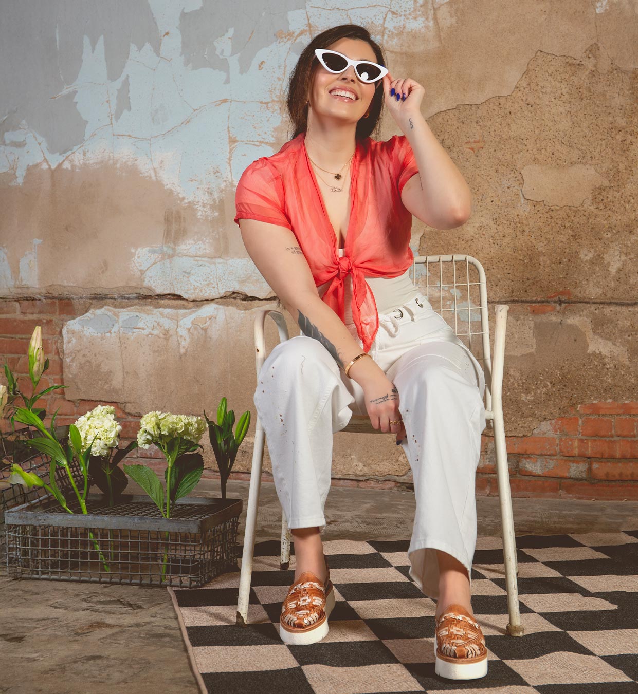 A person in white sunglasses, a red tied blouse, and white pants sits smiling on a metal chair. The scene is enhanced by the Lizzie platform huaraches from Bed Stu with hand-woven leather. A textured wall and potted plants on a checkered floor create an inviting ambiance.