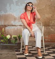 A person in white sunglasses, a red tied blouse, and white pants sits smiling on a metal chair. The scene is enhanced by the Lizzie platform huaraches from Bed Stu with hand-woven leather. A textured wall and potted plants on a checkered floor create an inviting ambiance.