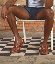 Someone sits cross-legged on a white chair, wearing dark shorts and Bed Stu Lone brown distressed leather sandals with an antique brass buckle, on a checkered rug by a brick wall.